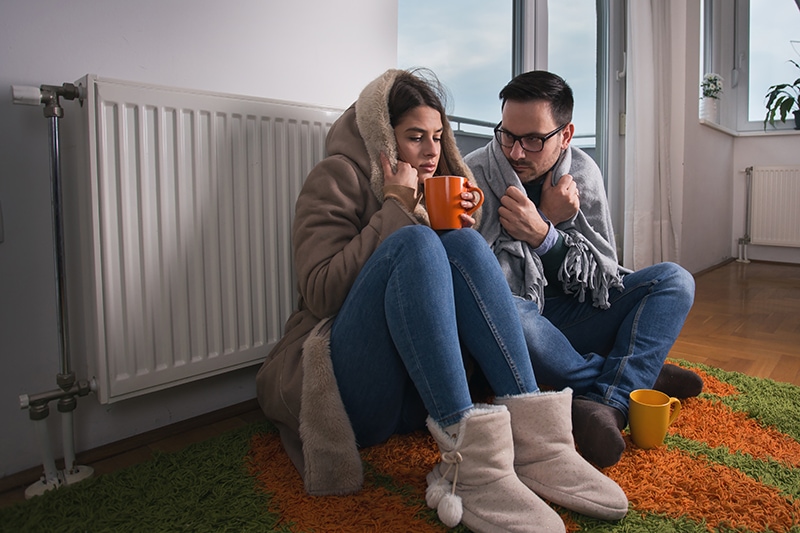 How Can I Make My Furnace Last Longer? Cold couple wrapped in blankets holding mugs next to furnace.