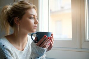 A woman with a coffee mug near a window. How to Lower Your Winter Heating Bill.